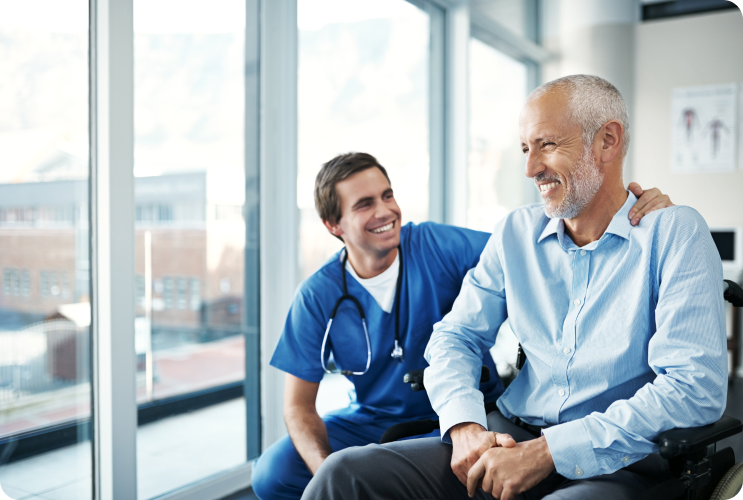 Healthcare professional smiling and reassuring an elderly male patient seated in a wheelchair inside a hospital room.