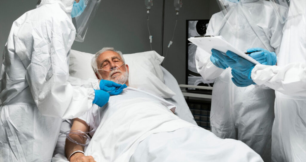 Medical team in protective suits attending to an elderly patient with a nasal cannula in a hospital bed.